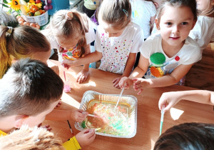 Dzieci stoją wokół stolika i zakraplają kolorowe mleko do kuwety z olejem
