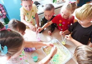Dzieci stoją wokół stolika i zakraplają kolorowe mleko do kuwety z olejem