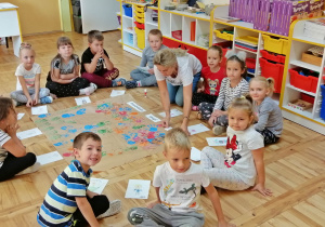 Dzieci siedzą z nauczycielem na podłodze wokół pracy plastycznej z odbitymi dłońmi