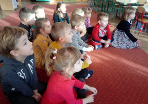 Dzieci siedzą na dywanie słuchając Mikołaja