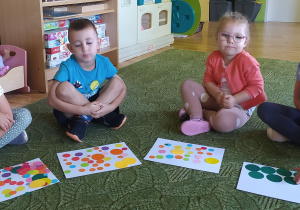 Dzieci siedzą na dywanie i prezentują ułożone na kartce kolorowe kropki
