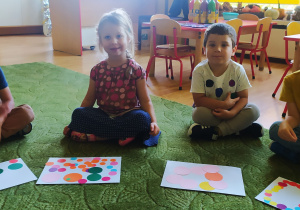 Dzieci siedzą na dywanie i prezentują swoje prace z kolorowymi kropkami