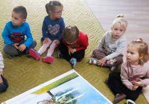 Dzieci siedzą na dywanie oglądając ilustrację drzewa