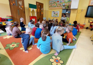 Dzieci siedzą na dywanie i słuchają książki czytanej przez nauczycielkę