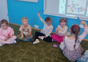 Dzieci siedzą na dywanie pokazując kolorowe kropki