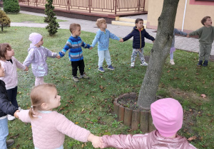 Dzieci stoją w ogrodzie trzymając się za ręce tworząc koło wokół drzewa